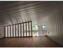 Dachgeschossraum mit schräger Holzdecke und holzverkleideten Wänden, Holzfußboden sowie einer niedrigen Trennwand mit senkrechten Streben. Große Fensterfront mit Blick auf Bäume; rechts ein kleiner Durchgang zu einem tieferen Bereich mit Treppe.