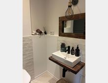 Kleines, modernes Badezimmer mit einer weißen Wand und weißen rechteckigen Fliesen. Ein Waschbecken mit schwarzer Armatur steht auf einer rustikalen Holzplatte, darüber hängt ein rechteckiger Spiegel mit einem Holzrahmen. Eine dekorative Hängelampe mit Metallkäfig und eingerollte Handtücher auf einer Ablage ergänzen das Design.