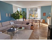 Wohnzimmer mit einer großen, beigen Eckcouch, zwei runden Couchtischen und einer Zimmerpflanze vor einer hellblauen Wand. Im Hintergrund Essbereich mit Holztisch, weißen Stühlen und großen Fenstern, durch die Tageslicht einfällt; heller Holzfußboden im Fischgrätenmuster.