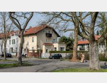 Straßenszene in einem Wohngebiet mit historischen Gebäuden, gekennzeichnet durch helle Fassaden und rote Schrägdächer. Im Vordergrund führen Kopfsteinpflaster, ein großes Paar Bäume und ein Fußgängerweg vorbei, während Menschen spazieren gehen und Autos am Straßenrand parken.