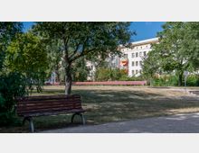 Städtischer Park mit Wiese, Bäumen und mehreren Parkbänken; im Hintergrund befinden sich mehrstöckige Wohngebäude mit hellen Fassaden und Balkonen.
