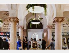 Innenraum eines historischen Gebäudes mit hohen Rundbögen und verzierten, sandfarbenen Säulen im korinthischen Stil. Menschen unterhalten sich in einer festlichen Atmosphäre, während hängende Pflanzen und moderne Leuchten die Decke schmücken; an den Wänden sind Schriftzüge erkennbar.