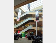 Einkaufszentrum mit mehreren Etagen, verbunden durch Rolltreppen. Im Vordergrund stehen Quads ausgestellt, während von der Decke Banner hängen, und Oberlichter lassen Tageslicht in den Innenbereich.