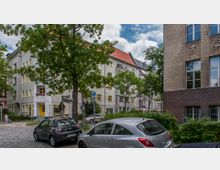 Straßenkreuzung in einem Wohngebiet mit Bäumen, parkenden Autos und mehrstöckigen Gebäuden mit hellen Fassaden und gelben Fensterrahmen. Im Vordergrund steht ein Richtungsschild, daneben Gehwege und Begrünung.