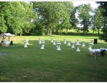 Eine Wiese mit verstreut aufgestellten Stehtischen, die mit weißen Hussen bedeckt sind, umgeben von hohen Bäumen. Im Vordergrund sind zwei Tische mit weißen Decken und Personen, die diese vorbereiten, sowie im Hintergrund eine landschaftliche Umgebung.