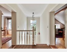 Flur eines Hauses im Obergeschoss mit einem weißen Holzgeländer und einem großen Fenster, das viel Tageslicht hereinlässt. Auf beiden Seiten des Flurs führen offene Türen in angrenzende Zimmer mit Holzböden und Teppichen.