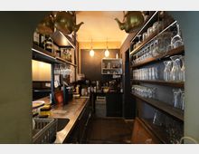 Blick in einen schmalen Bar- und Spülbereich mit Edelstahl-Arbeitsfläche, Spülbecken und Regalen mit Flaschen und hängenden Gläsern. Rechts stehen mehrere Holzregale voller Trinkgläser, im Hintergrund sind eine Kaffeemaschine und weitere Barutensilien unter warmen Deckenleuchten zu sehen.