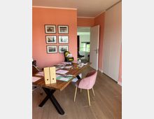 Ein Arbeitszimmer mit einer korallfarbenen Wand, an der sechs gerahmte Schwarz-Weiß-Fotografien hängen. Im Vordergrund steht ein Holztisch mit schwarzen Metallbeinen, der mit Papieren und Büromaterialien bedeckt ist. Links ein Bürostuhl, rechts ein rosa Polsterstuhl; im Hintergrund eine offene Tür mit Blick auf ein weiteres Zimmer.