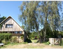 Ein rustikales, zweistöckiges Gebäude mit einer Holzfassade, großen Fenstern und einem mit Pflanzen bewachsenen unteren Teil. Es steht in einer ländlichen Umgebung mit großen Bäumen, einem Holzzaun und einer offenen Wiese unter klarem blauem Himmel.