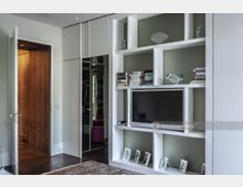 Ein modernes Wohnzimmer mit einem weißen Einbauregal, das einen Fernseher, Bücher, Deko-Objekte und Bilderrahmen enthält. Links befindet sich eine offene Tür aus Holz, daneben ein Schrank mit Spiegeltüren.