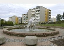 Ein kleiner Springbrunnen mit einer Tierfigur in der Mitte, umgeben von einer gepflasterten Fläche, vor mehreren mehrstöckigen Wohngebäuden mit farblich abgesetzten Balkonfassaden. Im Hintergrund befinden sich Grünflächen und Bäume.