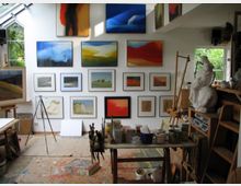 Ein Künstleratelier mit zahlreichen gerahmten Gemälden an einer weißen Wand, darunter abstrakte Landschaften in leuchtenden Farben. Im Vordergrund befinden sich Malutensilien wie Pinsel, Farben, eine Staffelei und eine Statue, während durch Fenster natürliches Licht hereinfällt.