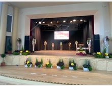 Eine Bühne in einem Veranstaltungssaal, dekoriert mit Blumenarrangements, Pflanzen und weißen Säulen mit buntem Blumenschmuck. Ein Banner mit der Aufschrift „Jugendweihe 2021“ hängt vor dem schwarzen Bühnenhintergrund, der mit Scheinwerfern beleuchtet ist.