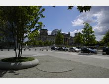 Blick auf das Reichstagsgebäude in Berlin mit seiner gläsernen Kuppel, umgeben von Bäumen und einem gepflasterten Platz. Im Vordergrund geparkte Autos und modern gestaltete Grünflächen.