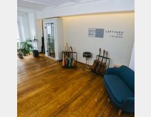 Ein heller Studio-Raum mit Holzboden und weiß gestrichenen Wänden. An der Wand stehen mehrere Gitarren in Ständern, eine Trommel und ein blaues Sofa; im Hintergrund befindet sich eine Glastür, daneben einige Topfpflanzen.