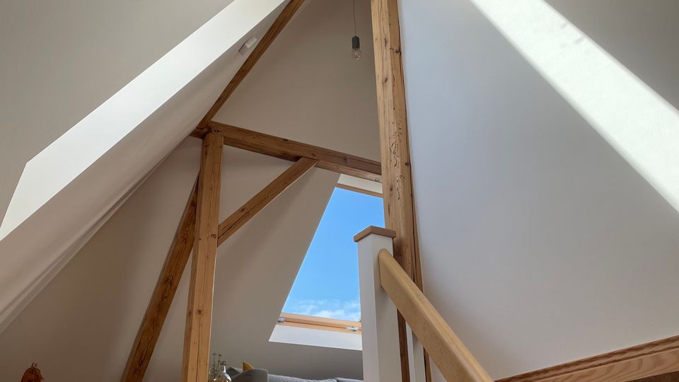 Blick vom Treppenaufgang in ein helles Dachgeschoss mit schrägen weißen Wänden und sichtbaren Holzbalken. Ein großes Dachfenster lässt Sonnenlicht auf eine kleine Sitzecke mit Sofa und Beistelltisch fallen.