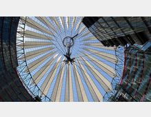 Blick nach oben auf das kreisförmige Dach des Sony Centers in Berlin, bestehend aus einer transparenten Stahl- und Glasstruktur mit weißen, segelartigen Membranen. Um das Dach herum sind moderne Glasfassaden mit roten Akzenten sichtbar.