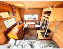 Innenraum eines Wohnwagens mit holzverkleideten Wänden und Decke. Links befinden sich eine Küchenzeile mit Schränken, einem Wasserkocher und Regalfläche, rechts ist eine Sitzecke mit Tisch und Stauraum, im Vordergrund ein Bett.