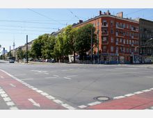 Straßenszene in einer städtischen Umgebung mit einer Kreuzung, einem Fahrradstreifen im Vordergrund und Straßenbahnleitungen in der Luft. Links sind Wohngebäude mit Bäumen im Hintergrund zu sehen, während rechts ein rotes Eckhaus mit Geschäften im Erdgeschoss steht.