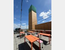 Rooftop terrace with orange tables, mixed seating, and string lights overhead, bordered by metal railings and a wood-paneled wall. A tall tower with tan cladding and a green glass top rises behind the patio, with city skyline views under a blue sky.