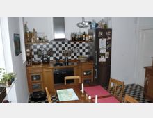 Küche mit Holzmöbeln, schwarzer und weißer Fliesenboden sowie schwarz-weiße Kachelrückwand. In der Mitte des Raumes steht ein Esstisch mit Holzstühlen, neben einem Kühlschrank mit Magneten an der Tür.