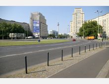 Straßenszene in einer Stadt mit breitem, geschwungenem Fahrbahnverlauf und Grünflächen in der Mitte; flankiert von großen, symmetrischen Gebäuden im Stil der Nachkriegsmoderne. Im Hintergrund ist der Berliner Fernsehturm sichtbar.