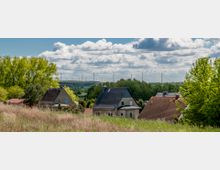 Ländliche Landschaft mit Häusern mit roten und dunklen Dächern, umgeben von Bäumen und Feldern; im Hintergrund sind Windräder und eine bewaldete Ebene zu sehen, unter einem Himmel mit teils bewölkten Abschnitten.