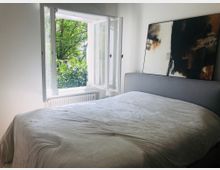 Schlafzimmer mit einem grauen Polsterbett und weißer Bettdecke, daneben ein abstraktes Gemälde an der Wand. Ein geöffnetes Fenster mit Ausblick auf grüne Bäume lässt Tageslicht in den Raum.