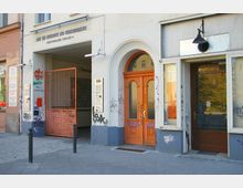 Fassade eines Gebäudes mit dem Schriftzug „Haus der Demokratie und Menschenrechte“ in der Greifswalder Straße 4. Zu sehen sind ein orangefarbenes Holztor, ein Durchgang mit einem Metalltor, Glaselemente sowie Graffiti an der Wand.