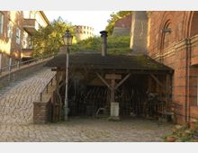 Ein gepflasterter Hof mit einem kleinen überdachten Holzunterstand, der von einem Geländer und dekorativen Metallzäunen umgeben ist. Im Hintergrund sind backsteinerne Gebäude, eine Laterne und ein Rundturm sichtbar, der von Grünflächen umgeben ist.