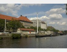 Ein Uferbereich mit Kanal, gesäumt von Gebäuden im klassischen und modernen Stil mit roten Ziegeldächern und hellen Fassaden. Im Vordergrund sind Anlegestellen, kleinere Boote und grüne Vegetation sichtbar, bei sonnigem Himmel mit weißen Wolken.