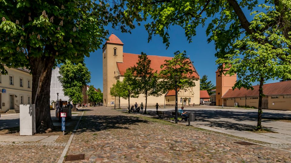 Gepflasterter Dorfplatz mit großen schattenspendenden Bäumen und Sitzbänken, umgeben von niedrigen Gebäuden. Im Hintergrund steht eine helle Kirche mit rotem Satteldach und Turm unter blauem Himmel.