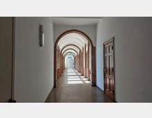 Langer, heller Flur mit gemauerten Rundbögen aus roten Ziegelsteinen auf der linken Seite und einer geschlossenen Holztür rechts; am Ende des Flurs befinden sich eine Glastür und mehrere Schilder.