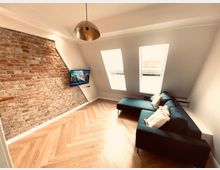 Wohnzimmer in einer Dachgeschosswohnung mit sichtbarer Backsteinwand, einem schrägen Dachfensterbereich mit zwei Fenstern und einer schwarzen Eckcouch mit hellen Kissen. Ein Flachbildfernseher ist an der Backsteinwand montiert, und der Boden besteht aus hellen Holzdielen im Fischgrätmuster.