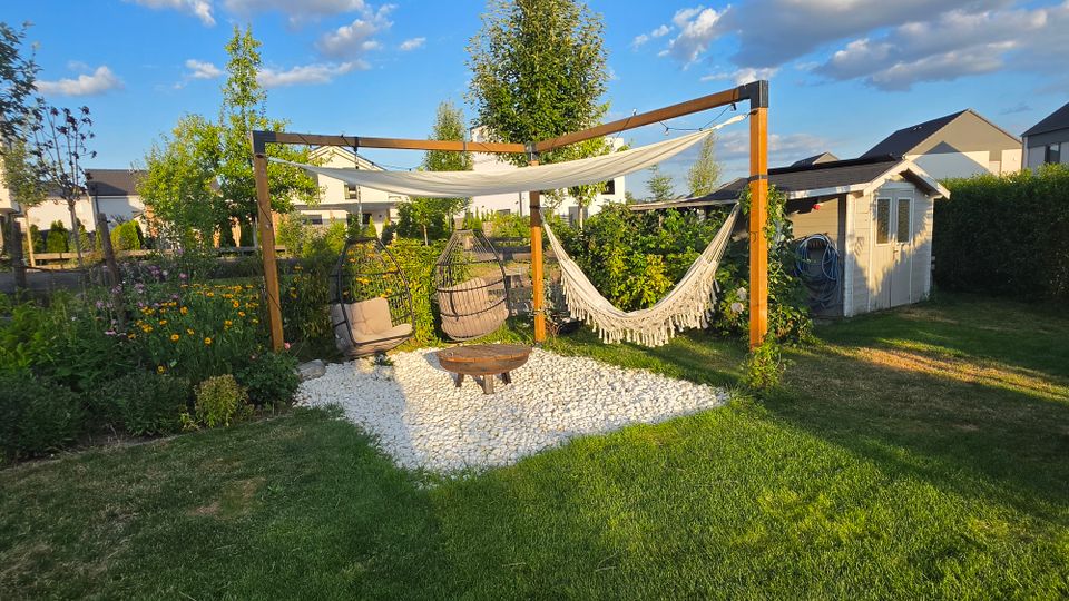 Garten-Lounge-Bereich mit Holzpergola, darüber einem Sonnensegel und seitlich einer Hängematte, auf einer Fläche aus weißen Kieseln mit Feuerstelle und Hängesesseln. Im Hintergrund sind Blumenbeete, Rasen, ein kleines Gartenhaus und Wohnhäuser zu sehen.