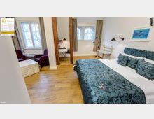 Helles Hotelzimmer mit Holzboden, einem Doppelbett mit gemustertem Überwurf in Blau und Grün sowie passenden Kissen. Im Raum befinden sich außerdem ein kleiner Schreibtisch mit Stuhl, zwei Sessel in einer Sitzecke und zwei große Fenster mit Vorhängen, die Tageslicht hereinlassen.