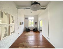 Heller Raum mit Holzdielenboden, weißen Wänden und einer großen hohen Fensterfront im Hintergrund. An der linken Wand hängen gerahmte Kunstwerke, während zwei schwarze Ledersessel vor dem Fenster stehen; eine Tür rechts führt in einen angrenzenden Raum.