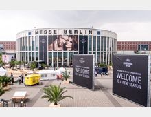 Runde Veranstaltungshalle „Messe Berlin“ mit großem Schriftzug auf der Fassade und Werbeplakaten für „Panorama Berlin“ darüber. Im Vordergrund ein gepflasterter Platz mit Palmen, weißen Zelten und einem gelben Verkaufswagen.