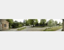 Pflasterstraße in einem ländlichen Dorf mit alten Gebäuden, darunter ein rotes Backsteinhaus links und ein grünes Haus rechts. Die Straße ist von Bäumen und einer Steinmauer gesäumt, im Hintergrund ist eine ruhige Grünfläche zu sehen.