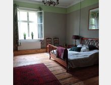 Ein Schlafzimmer mit Holzdielenboden, einem Bett mit Holzkopfteil, bunten Kissen und einer grau-beigen Tagesdecke. Im Raum befinden sich ein geöffnetes Fenster mit Vorhängen, zwei Stühle, ein Nachttisch mit Lampe und ein roter Teppich.