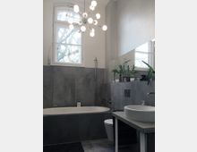 Modernes Badezimmer mit grauen Fliesenwänden und -boden, einer Badewanne, einem runden Waschbecken auf einer Ablage sowie einer Toilette. Ein großes Fenster mit Blick auf Bäume und eine auffällige Deckenleuchte runden die Einrichtung ab.