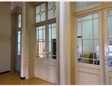 Holzrahmen-Türen mit großen Fensterscheiben in einem hellen Innenraum, flankiert von cremefarbenen Wandpaneelen. Im Hintergrund erkennbar: ein weiterer Raum mit gelben und weißen Wänden sowie Möbeln.