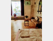 Ein gemütlich eingerichteter Raum mit Holzdielenboden, einer antiken Polstercouch, einem kleinen Holztisch mit einem Blumenstrauß, einem Stuhl vor einem großen Fenster mit tiefroten Vorhängen und einer sichtbaren Heizung. Die Wände sind in einem abgenutzten, teilweise freiliegenden Zustand, und ein gefaltetes Handtuch liegt auf einem Tuch im Vordergrund.