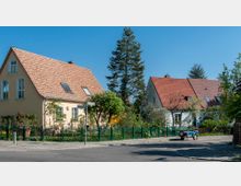 Straßenszene mit zwei Einfamilienhäusern mit roten Ziegeldächern und hellen Fassaden, umgeben von Gärten mit Bäumen und Blumen; vor den Gärten verläuft ein grüner Metallzaun, und ein kleiner Anhänger steht am Straßenrand.