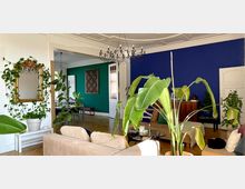 Wohnzimmer mit Pflanzen und auffälligen Wandfarben: Eine blau gestrichene Wand mit einem Holzmöbel und weißen Türen sowie eine grüne Wand mit einem dekorativen Wandobjekt. Im Raum befinden sich zahlreiche große Zimmerpflanzen, ein beiger Sessel und eine goldgerahmte Wandspiegeldekoration. Ein Kronleuchter hängt von der reich verzierten Decke.