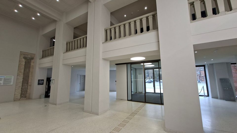 Helle, hohe Eingangshalle mit großen weißen Pfeilern, umlaufender Galerie mit Balustrade und Deckenleuchten. Vorne befinden sich automatische Glastüren zum Außenbereich sowie ein taktiles Leitsystem im hellen Steinboden.