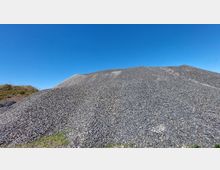Ein großer Hügel aus Schottersteinen unter klarem, blauem Himmel. Am unteren Rand sind vereinzelte grüne Grasbüschel sichtbar.