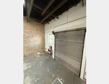 Innenraum einer hohen Lager- oder Werkstatthalle mit sichtbaren Deckenbalken, Backsteinwand und grauem Betonboden. Rechts befindet sich ein großes metallisches Rolltor neben einem kleinen Fenster in einer weißen Wand.