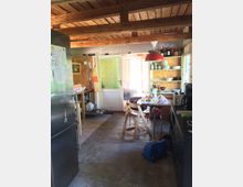 Rustikale Küche mit Holzdecke und sichtbaren Balken. Im Raum stehen ein Kühlschrank, ein Tisch mit Stühlen und offene Regale mit Geschirr; rechts ist ein dunkler Schrank, links eine Tür mit Glasfenster, die zu einem helleren Raum führt.