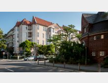Straßenszene in einer Wohngegend mit historischen Gebäuden: Links ein mehrstöckiges Wohnhaus mit hellem Putz und roten Dächern, rechts ein rundes Backsteingebäude mit Schieferdach. Im Vordergrund eine befahrene Straße, Fußwege und Bäume.